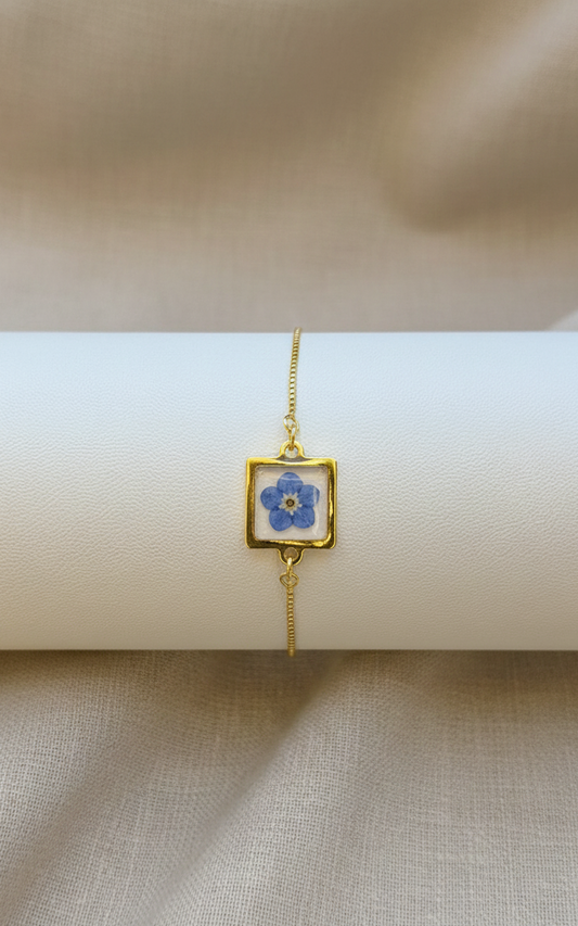 Gold bracelet with a square charm featuring a blue flower design on a beige surface.