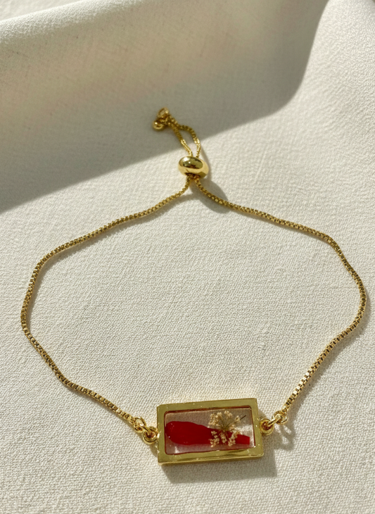 Gold bracelet with a rectangular charm featuring red and gold elements on a light fabric background